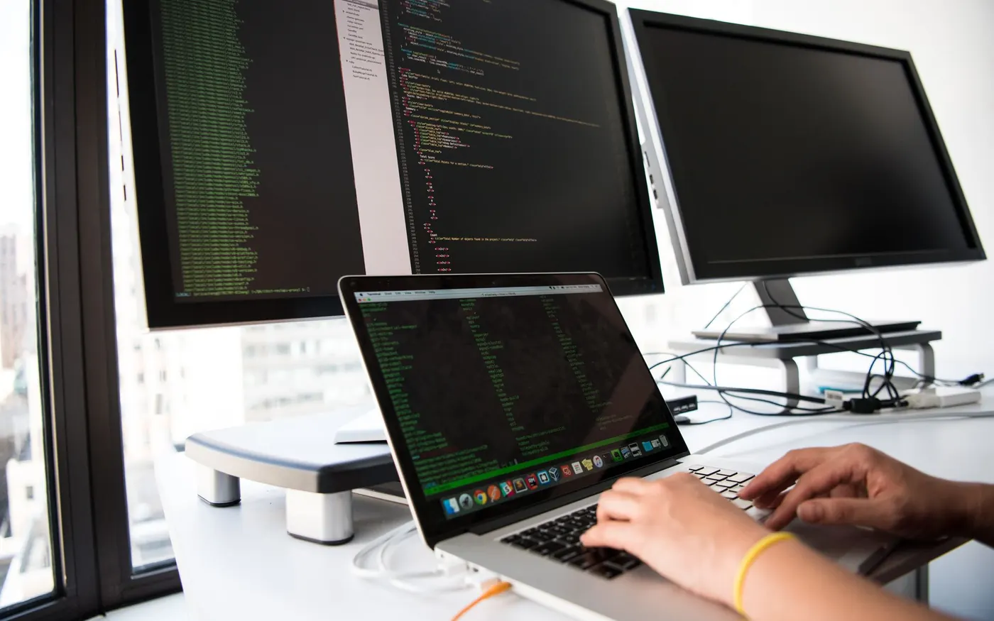 Hands typing code on a laptop in front of two large monitors showing more code