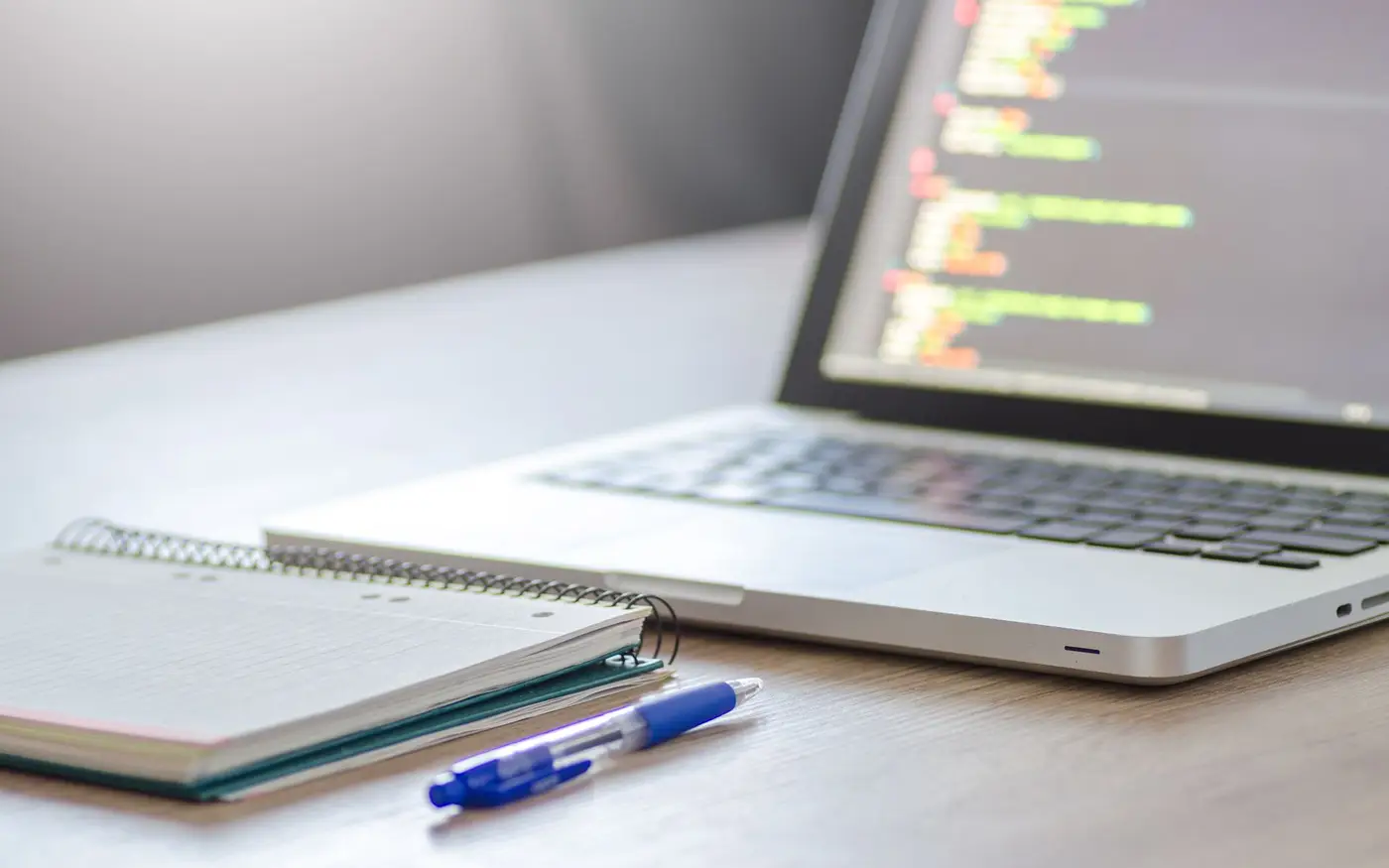 A laptop showing code beside an open notebook and a pen on a studio desk