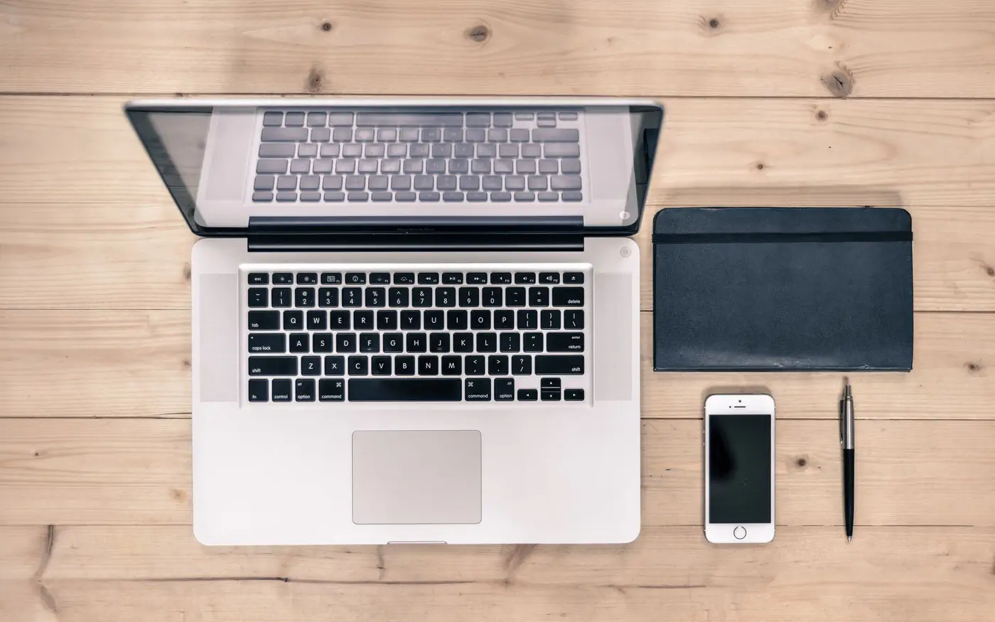 A laptop, phone, notebook and pen laid out on a wooden desk, seen from above